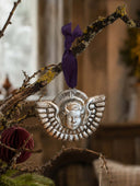 A silver-toned metal angel ornament with a purple ribbon hanging from a rustic tree branch in a softly blurred indoor setting.