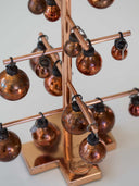 A copper-colored metal tree ornament display stand with multiple small hanging copper Christmas baubles, shown close up against a light neutral background.