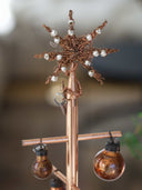 A copper-toned decorative tree stand with a star topper and hanging bulb ornaments, photographed against a softly blurred indoor background.