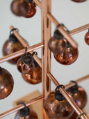 Close-up of a copper-toned geometric wall decor piece with hanging metallic glass ornaments on a light neutral background.