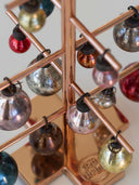 A close-up of a copper-colored geometric holiday ornament tree stand holding multiple reflective glass Christmas baubles in silver, gold, red, blue, and bronze tones.