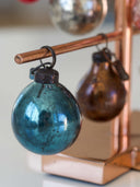 A close-up of a decorative hanging ornament set on a copper-colored display stand. The front ornament is a large, metallic teal-blue glass bauble with a dark cap, while a second amber-brown glass ornament hangs behind it on a rod.