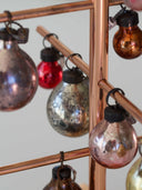 A close-up of a copper-colored metal hanging ornament rack with multiple vintage-style mirrored glass bulbs in silver, red, bronze, and pink tones.