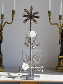 A silver metal jewelry display tree styled like a Christmas tree, holding silver crescent moon and star pendants, beaded necklaces, and two white fabric flowers, set on a marble mantel between two gold candelabras.