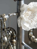 Close-up of a silver-toned jewelry display stand holding beaded necklaces and earrings, with a large white paper flower attached near a white peony flower, against a soft gray background.