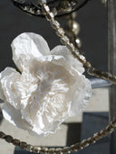 A close-up of a large white paper flower with layered ruffled petals, hanging near a beaded chain against a dark gray background.