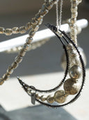 A close-up of a beaded crescent moon necklace with metallic silver beads, textured black wire, and a small silver bird charm, hanging against a softly blurred light gray background.