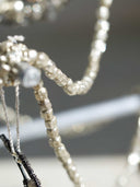 Close-up of a beaded necklace with clear faceted beads and a black braided cord detail, photographed against a soft blurred gray background.