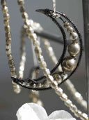 A close-up of a silver crescent moon pendant necklace with textured metallic beads and dark wrapped wire details, hanging against a soft gray background.