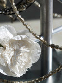 A close-up of a white textured flower-like decorative piece with layered ruffled petals, overlaid by strands of faceted metallic beads in silver and dark bronze tones against a gray industrial background.