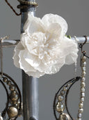 A large white paper flower with layered, ruffled petals hanging from a silver metal display stand, surrounded by draped beaded jewelry strands against a plain gray background.