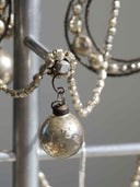 A close-up of a silver-toned beaded necklace with a hanging vintage-style silver ornament pendant, displayed on a metallic stand against a soft gray background.