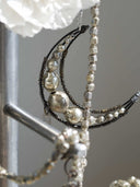 A close-up of a silver crescent moon necklace with textured beads and faceted crystal details, hanging on a display stand.